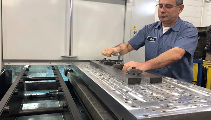 Machinist wearing blue shirt and safety glasses clamping parts onto a MIDACO Pallet Changer aluminum pallet shuttle system mounted outside the vertical machining center in a machine shop.